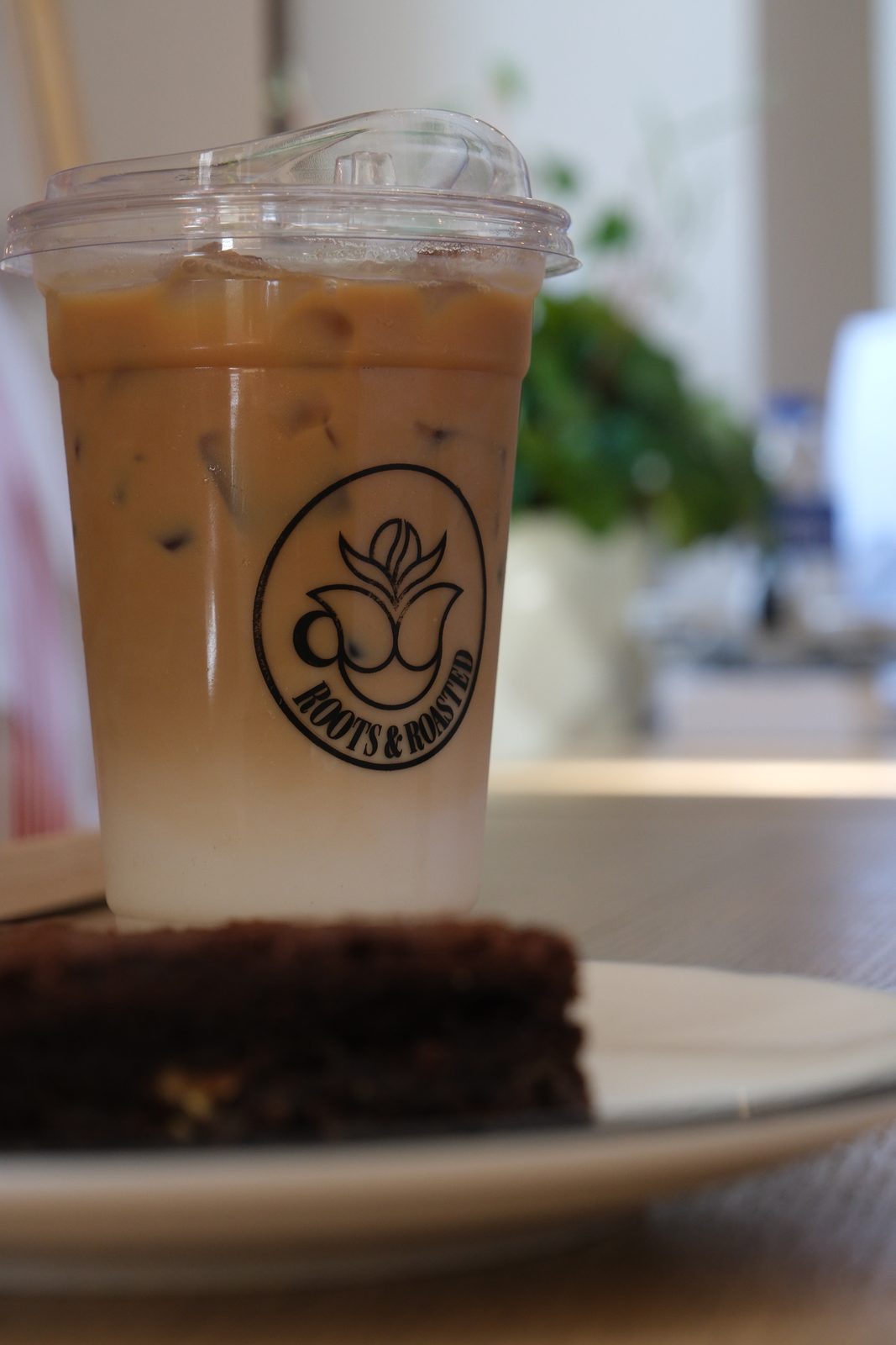 Iced latte in a branded Roots & Roasted cup beside a slice of cake